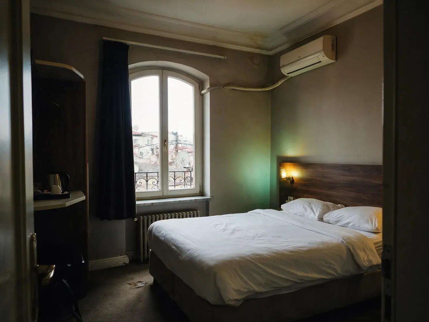 Before and after of a guest room in a converted karaköy han shot on a phone, with the window blown to white and the rest of the room sunk in shadow — original listing photo before editing
