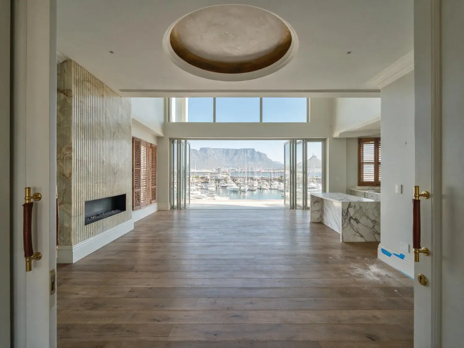 Before and after of an empty penthouse at one&only cape town is staged into a safari-lux salon, framing the existing table mountain and marina beyond — original listing photo before editing