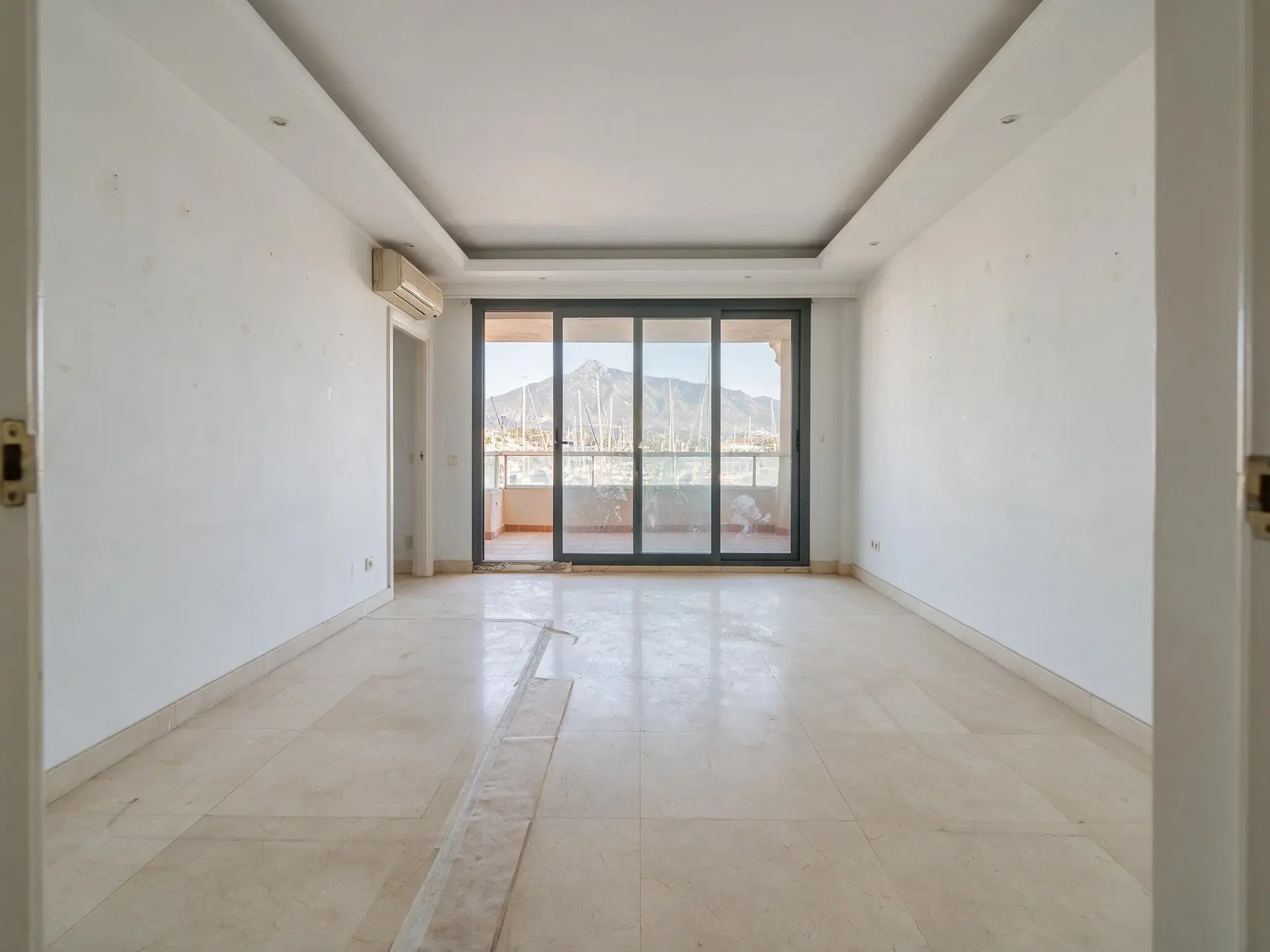 Before and after of an empty apartment salon overlooking Puerto Banús marina in Marbella, furnished by virtual staging. — original listing photo before editing