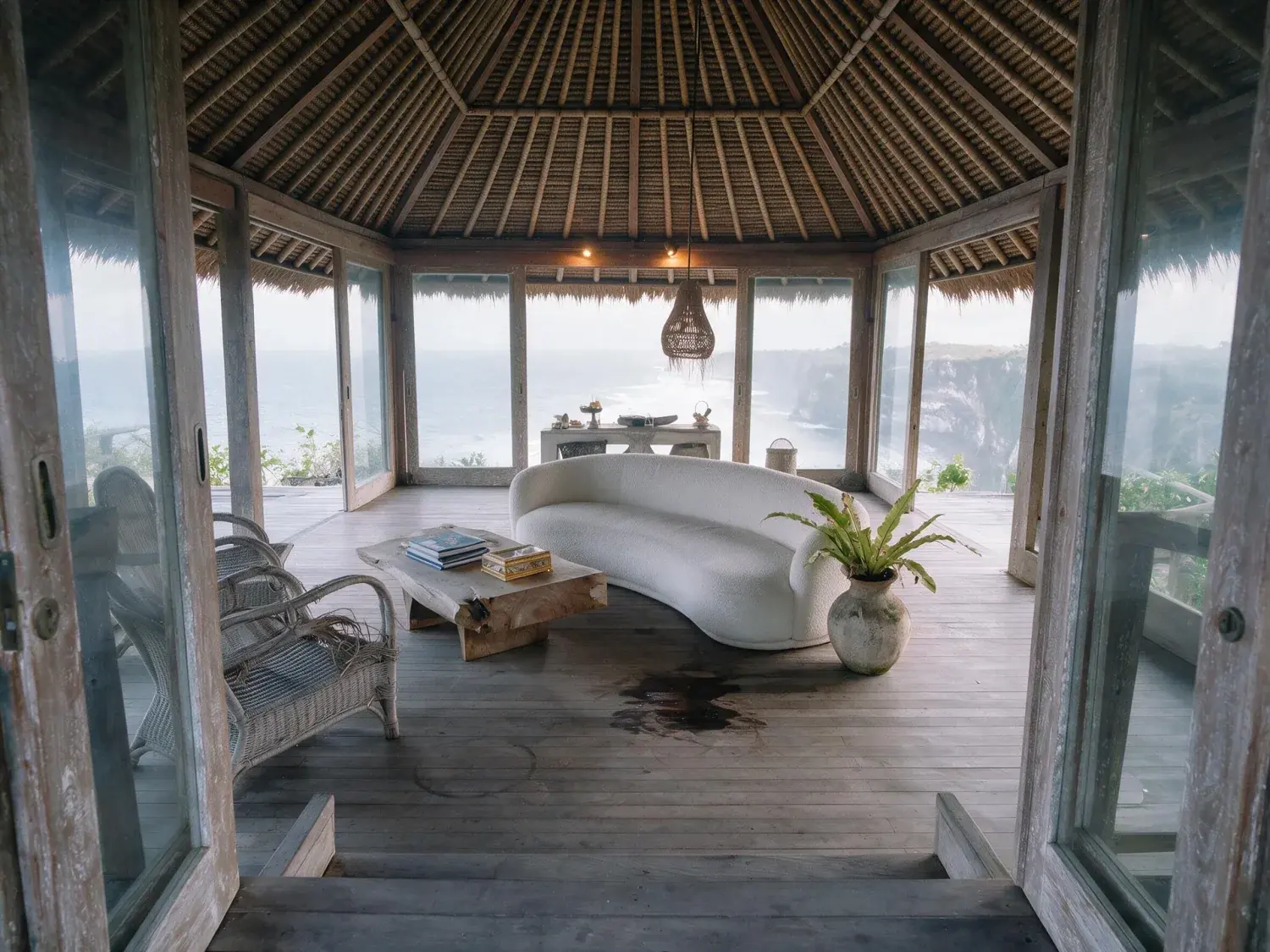 Before and after of a bali bukit cliff-villa pavilion lifts from a dim tropical-haze underexposure into full teak, rattan and indian ocean clarity — enhanced by Plotpane