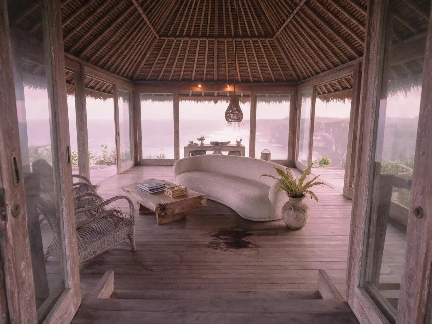 Before and after of a bali bukit cliff-villa pavilion lifts from a dim tropical-haze underexposure into full teak, rattan and indian ocean clarity — original listing photo before editing