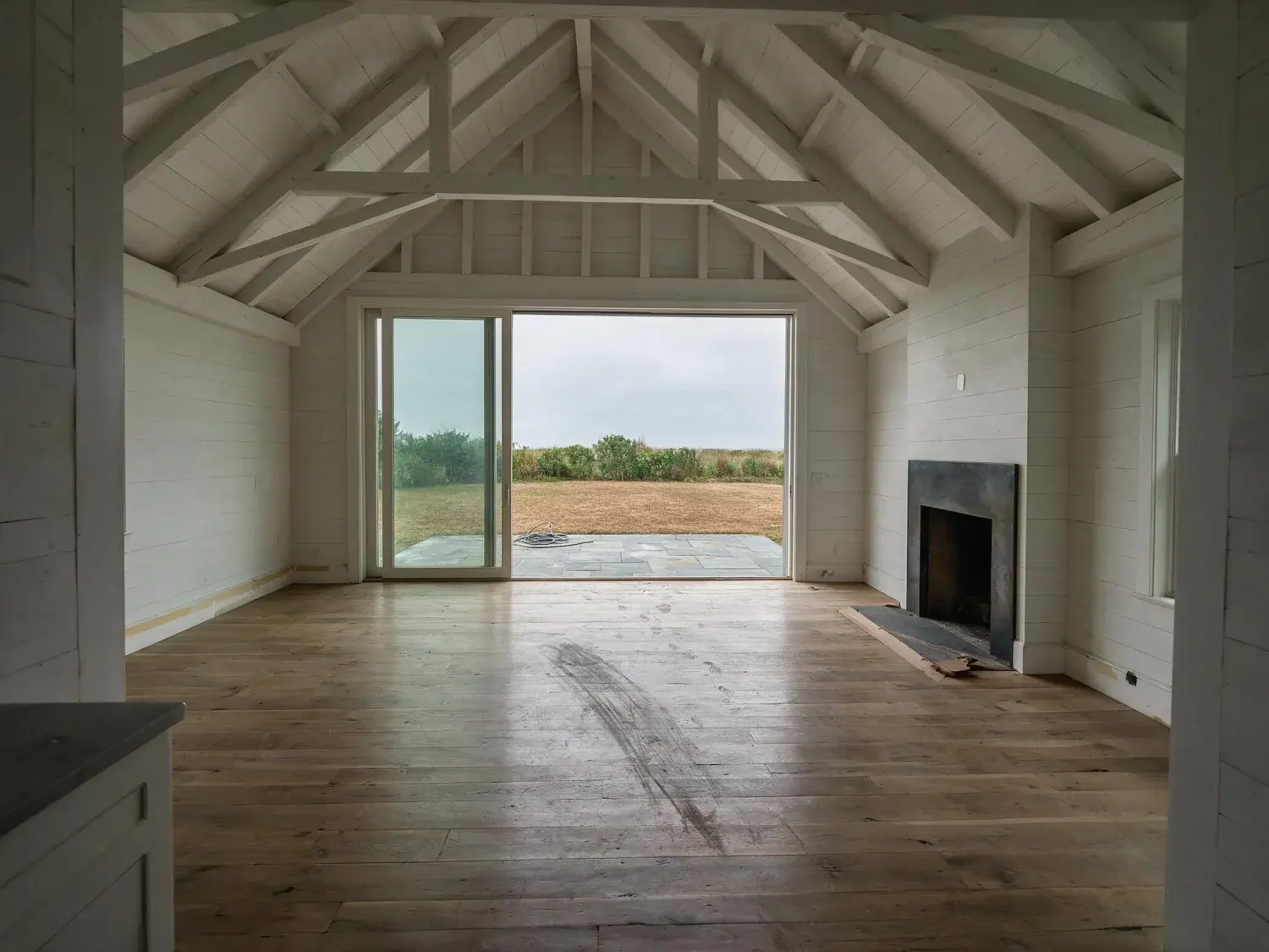 Before and after of a $12m bridgehampton modern-barn estate where virtual staging ai could stage the living room but couldn't fix the gray sky, brown lawn, or the hose coiled on the patio — original listing photo before editing