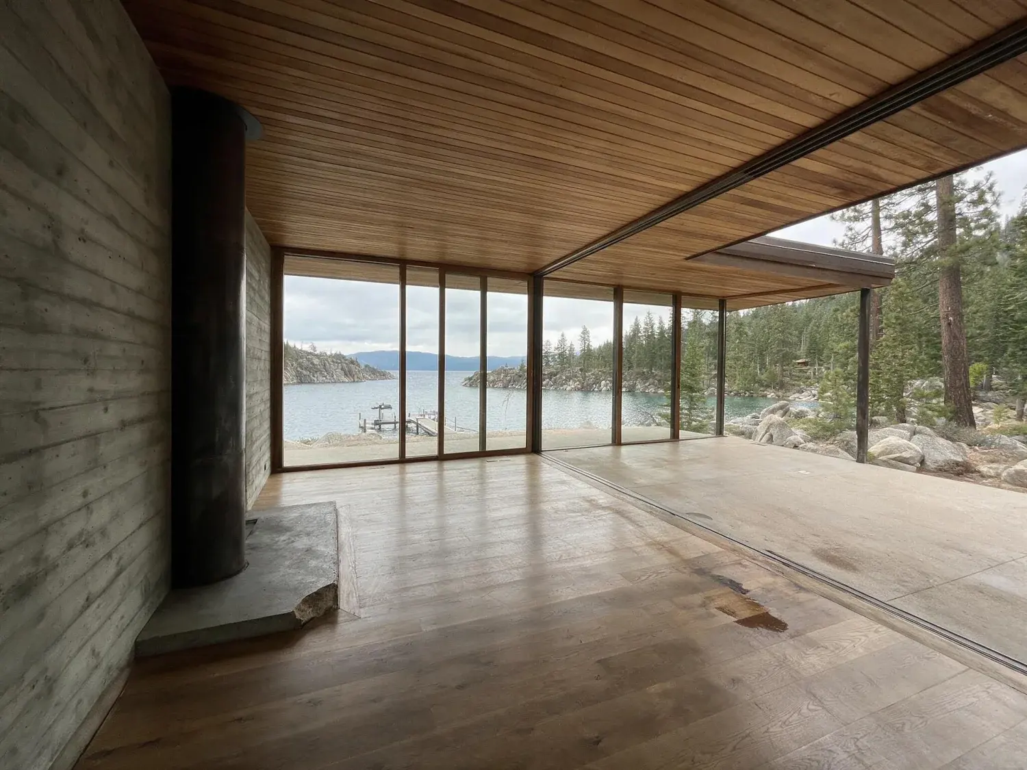 Before and after of a lake tahoe modernist retreat photographed from the lake side at golden hour — douglas fir forest, granite cliffs, alpine water framing a low-slung contemporary home — original listing photo before editing