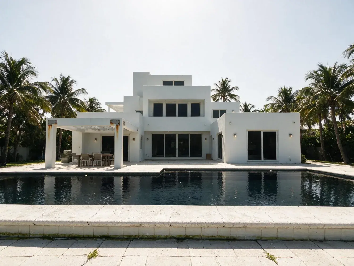 Before and after of a sculptural miami waterfront villa trades flat afternoon glare for a neon-drenched florida blue hour with every window ablaze — original listing photo before editing