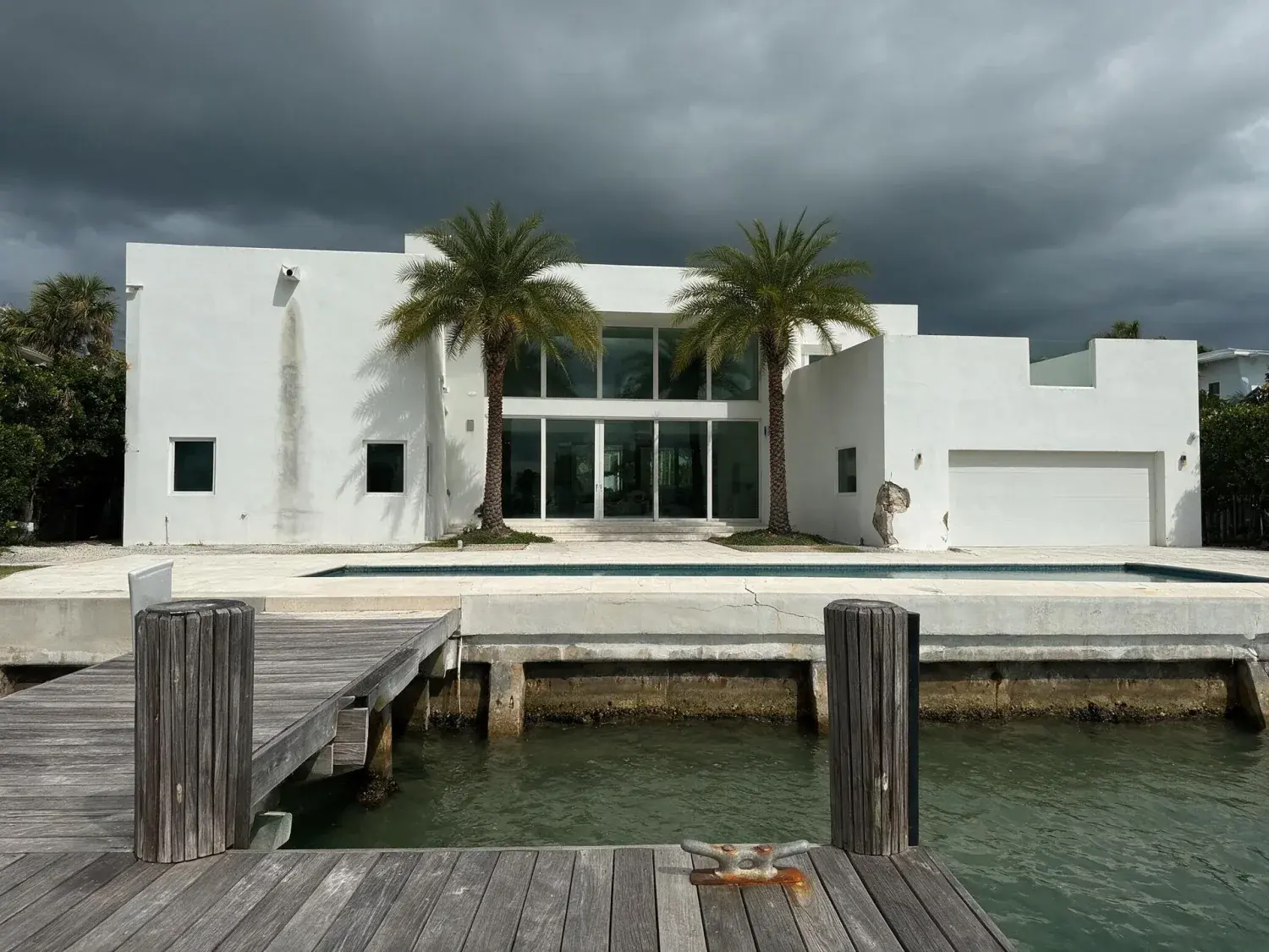 Before and after of a white-stucco miami waterfront modern villa lifted from a stormy grey sky into a postcard tropical azure — original listing photo before editing