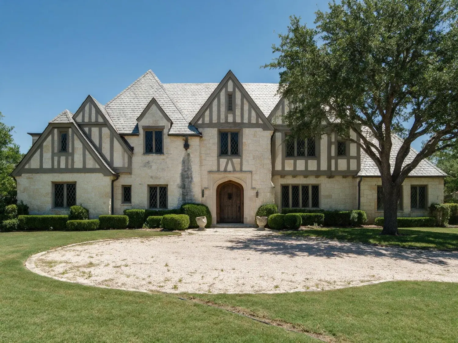 Before and after of front exterior of a highland park limestone-and-slate tudor estate converted from harsh midday to warm texas twilight — original listing photo before editing