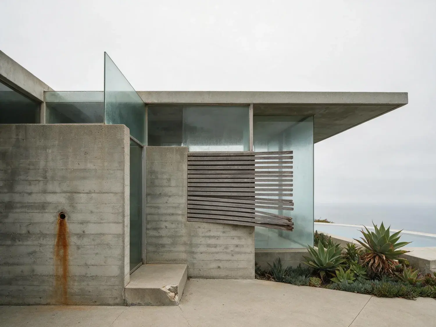 Before and after of a minimalist board-formed-concrete malibu retreat freed from a dull marine-layer sky and given a pacific-blue dreamscape — original listing photo before editing