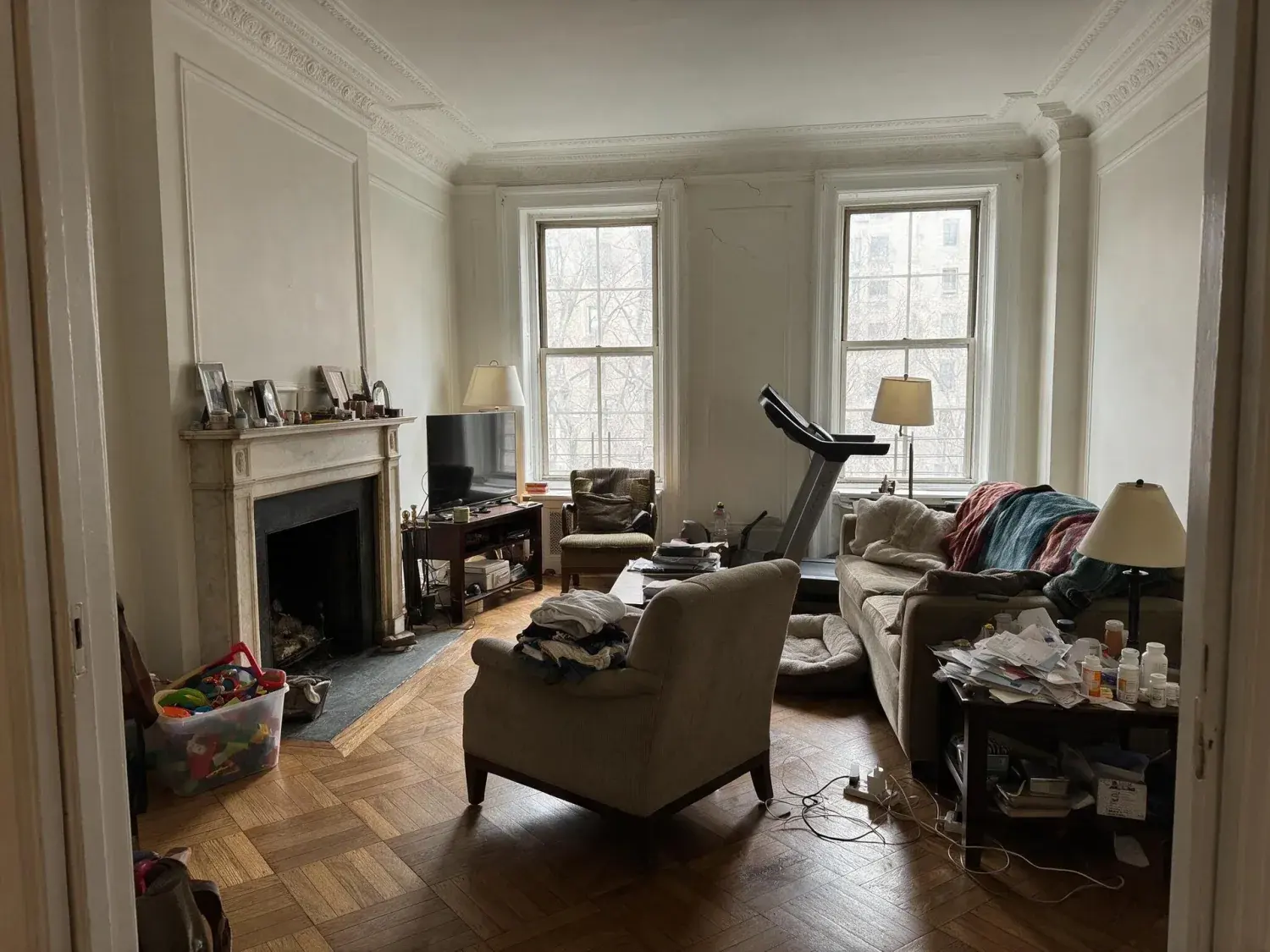 Before and after of a grand prewar park avenue living room with crown moldings and herringbone floors, stripped of a family's lived-in clutter to reveal its bones — original listing photo before editing
