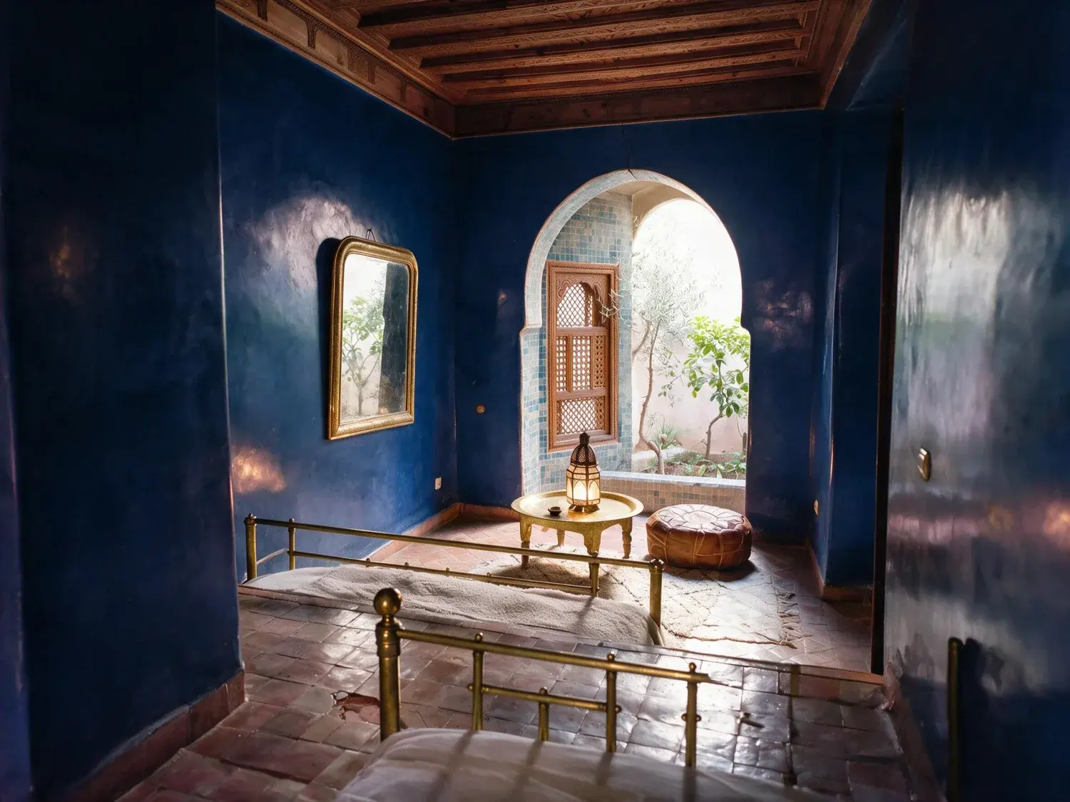 Before and after of a jardin-majorelle-adjacent boutique riad bedroom suite resolves from a muddy underexposure into full zellige and tadelakt colour — distinct from the courtyard riad already in the library — enhanced by Plotpane