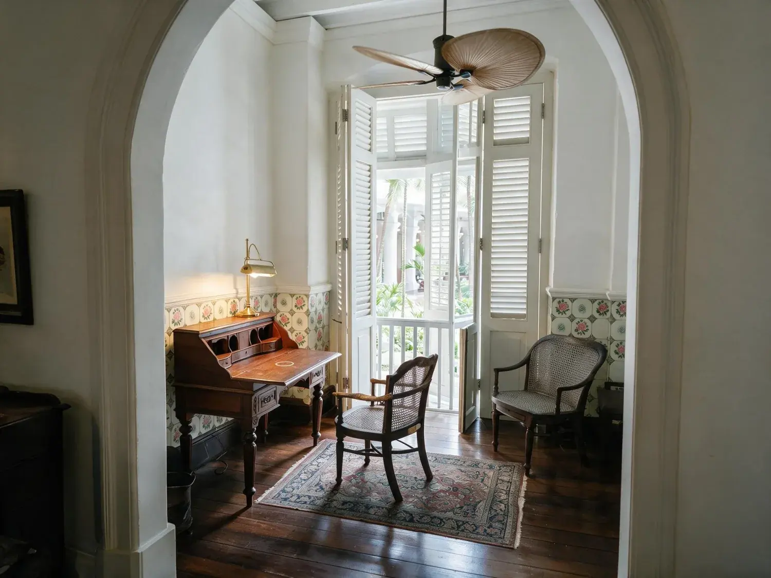 Before and after of a raffles singapore heritage suite writing room emerges from a muddy underexposed capture into crisp colonial clarity — teak, rattan, and courtyard palms — enhanced by Plotpane