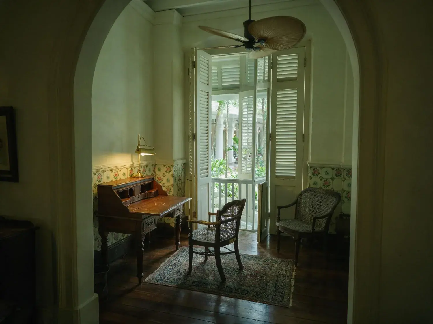 Before and after of a raffles singapore heritage suite writing room emerges from a muddy underexposed capture into crisp colonial clarity — teak, rattan, and courtyard palms — original listing photo before editing