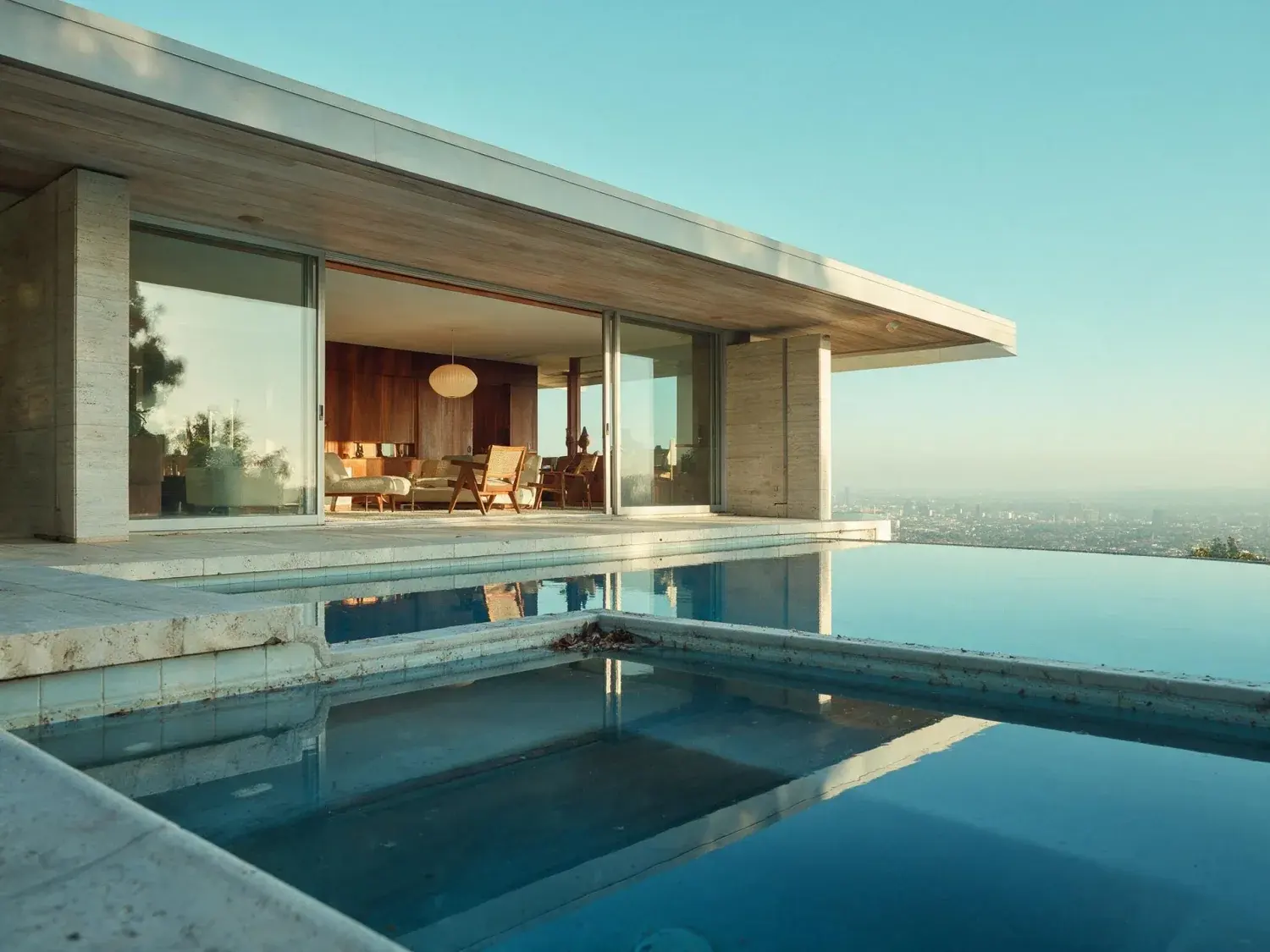 Before and after of a low-slung trousdale modernist glows from within at dusk, its cantilevered roof floating over an infinity pool poised above the la basin — original listing photo before editing