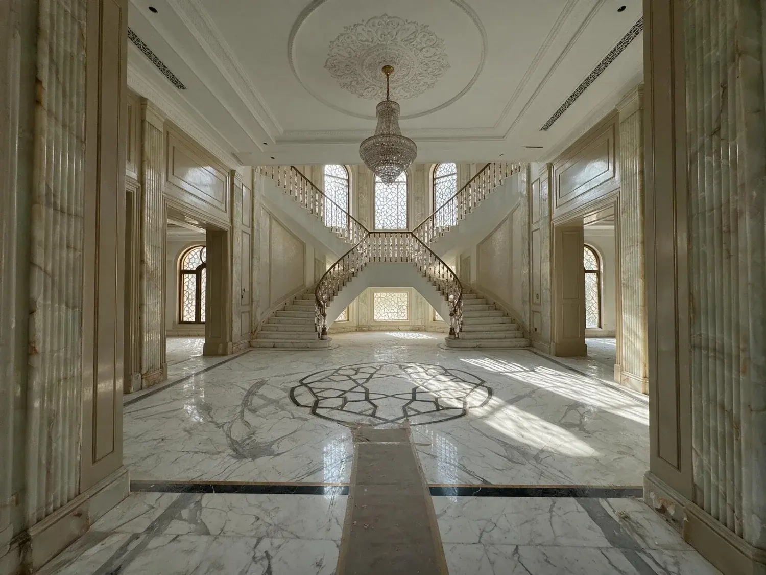 Before and after of an empty triple-height majlis hall in an emirates hills mansion awaits its furniture, its marble floor a silent mirror for a crystal chandelier — original listing photo before editing
