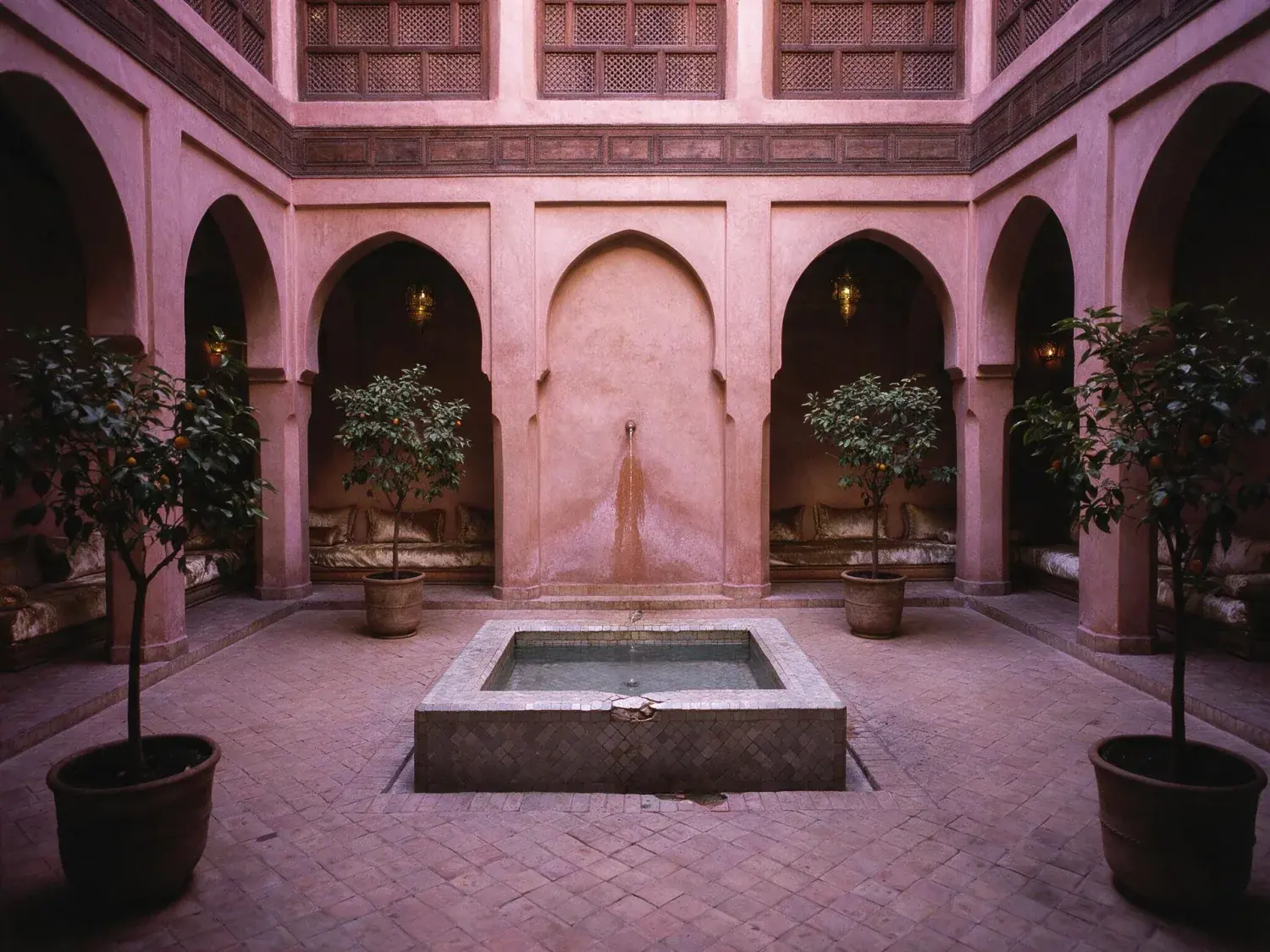 Before and after of a jewel-box marrakech riad courtyard recovers its tile colour and shadow depth, zellige glowing and orange leaves finally readable — original listing photo before editing