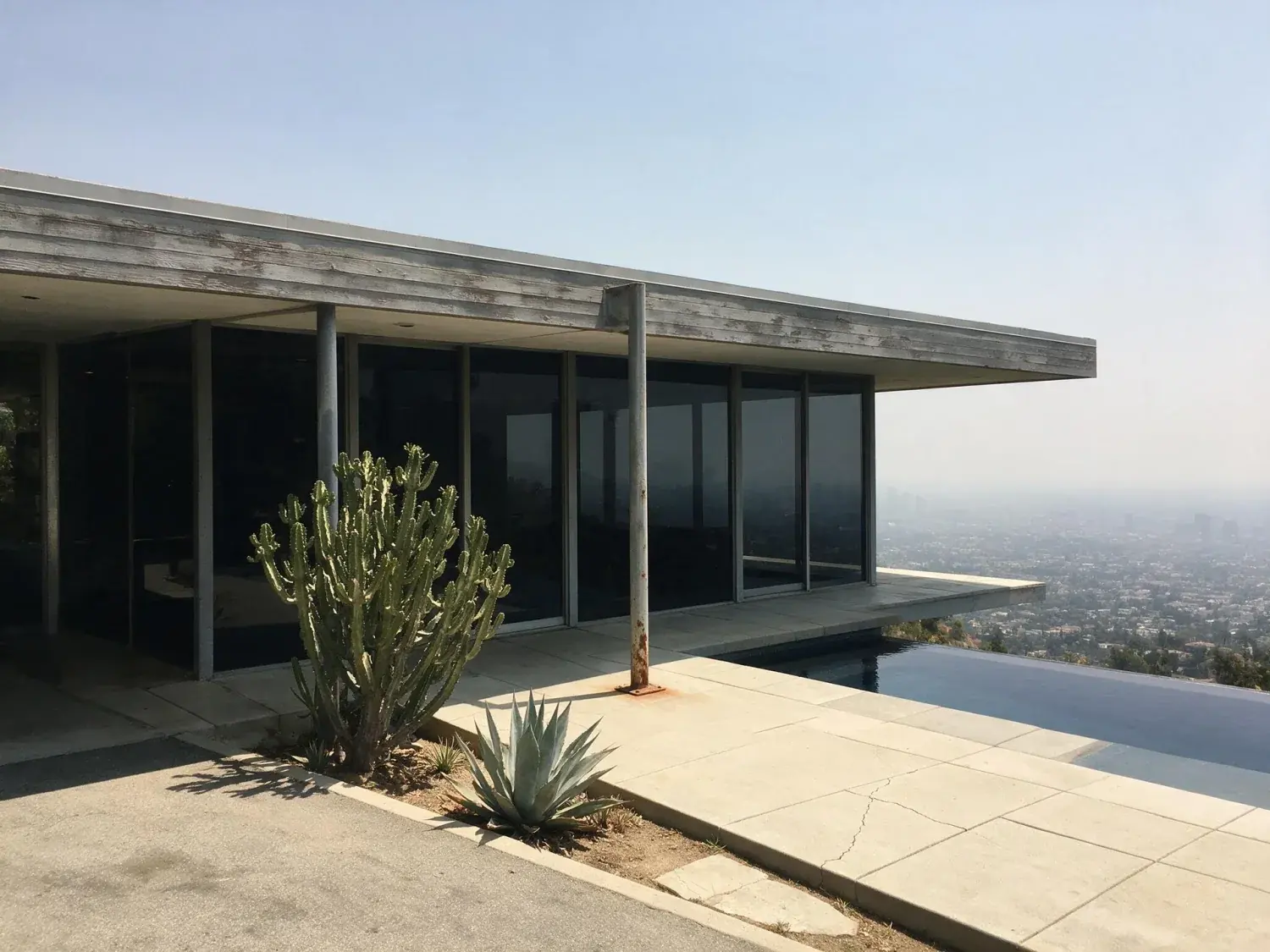 Before and after of a neutra-inflected bird streets cantilever spreads over the city-lights carpet below — flipped from hard midday glare to a slim aarons-register twilight — original listing photo before editing