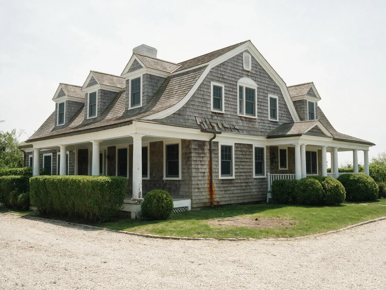 Before and after of a weathered-cedar hamptons estate with wraparound porch and manicured privet hedges, transformed from flat noon to a magazine-cover blue-hour portrait — original listing photo before editing