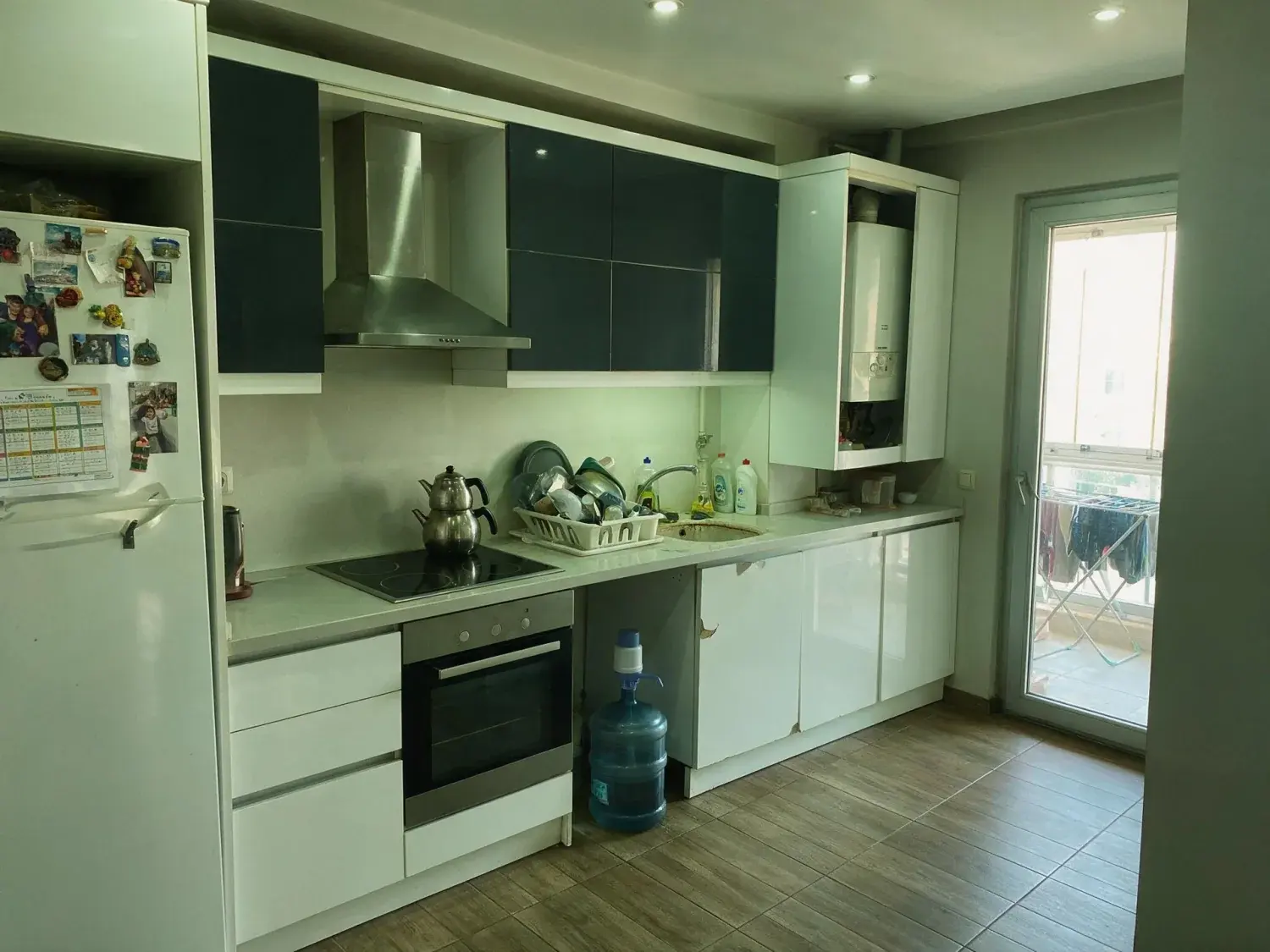 Before and after of a lived-in kitchen in an ataşehir residence tower, with the day's dishes, bottles and water carboy named for removal — original listing photo before editing
