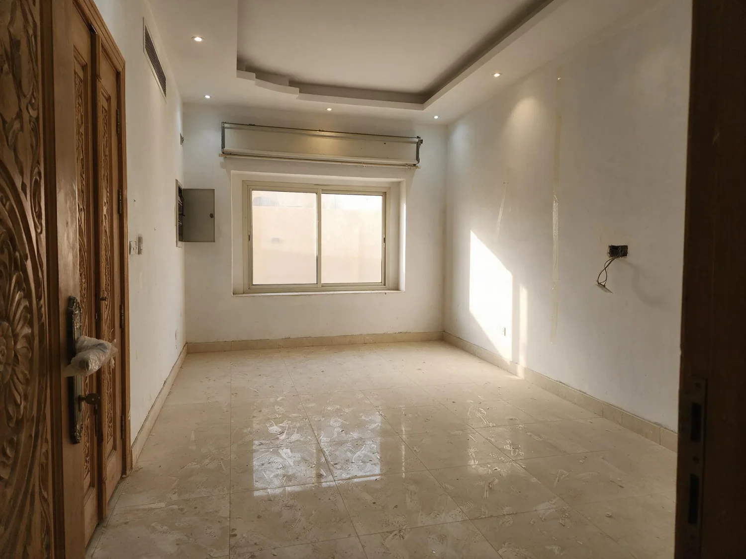 Before virtual staging: empty villa majlis with a carved wooden door, marble floor, AC grille and a loose wire on the wall
