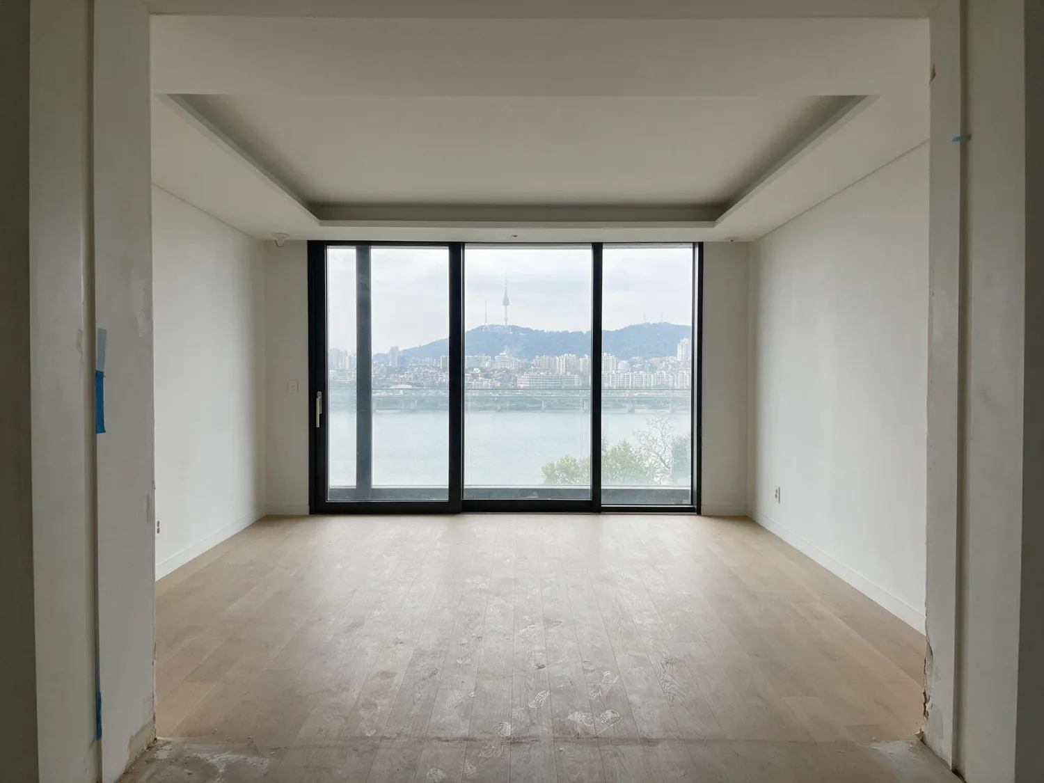 Before virtual staging: empty river-view living room with a full glass wall, oak floor and paint spatter near the doorway